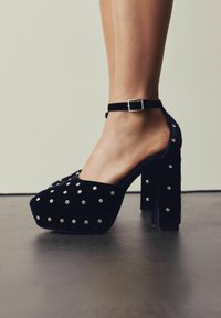 Juodas zomšinis aukšto kulno platformos batelis su sidabriniais kalnų kristalais, kulkšnies dirželiu ir sagtimi, dėvimas ant kojos, stovinčios ant tamsaus grindų paviršiaus.