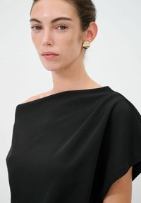 Abito nero off-the-shoulder in tessuto morbido, con design drappeggiato e scollo rotondo; abbinato a orecchini d'oro a cerchio.