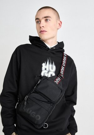 Młody mężczyzna w czarnej bluzie z kapturem z logo Hugo oraz czarnej torbie na ramię z logo Hugo na pasku i kieszeni.