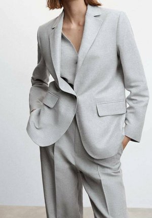 Vrouw draagt een lichtgrijs op maat gemaakt pak met een blazer over bijpassende broek, handen in de zakken, staand tegen een effen achtergrond.