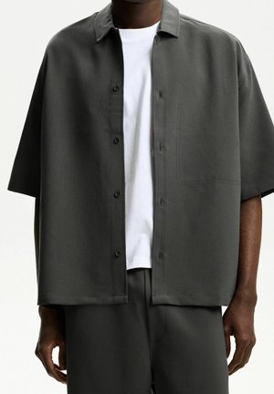 Mężczyzna ubrany w ciemnoszarą, krótkorękawową koszulę na guziki na białą koszulkę, w dopasowanych ciemnoszarych spodniach, stojący na białym tle.