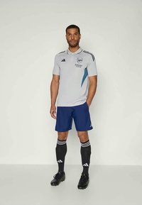 Hallikas Adidas jalgpallipluus siniste aktsentidega, eksponeeritud siniste lühikeste pükste, mustade sokkide, millel on valged triibud, ja mustade spordijalatsitega.