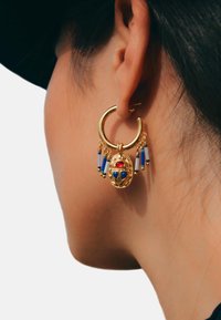 Boucle d'oreille en or avec un motif central de scarabée, orné d'accents rouges et bleus. Des franges avec des perles bleues pendent en dessous pour un détail supplémentaire.