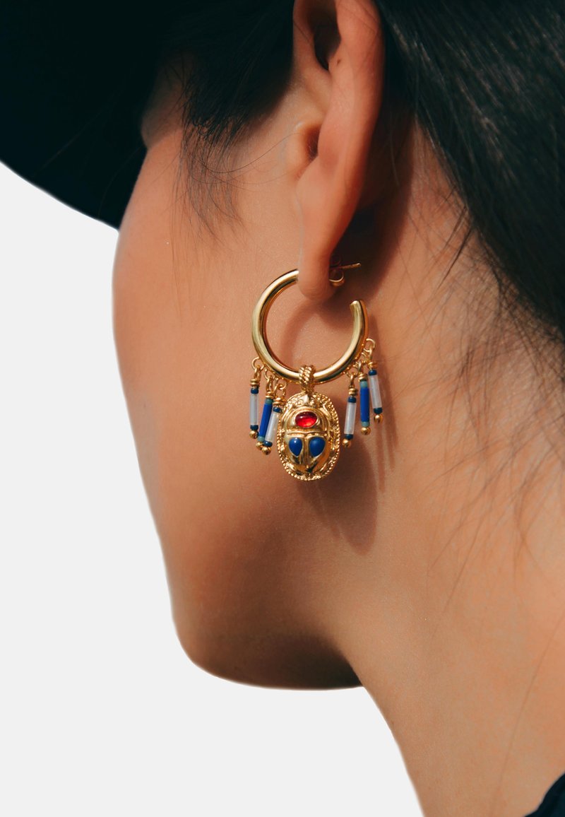 Boucle d'oreille en or avec un motif central de scarabée, orné d'accents rouges et bleus. Des franges avec des perles bleues pendent en dessous pour un détail supplémentaire.