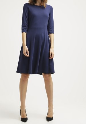 Femme portant une robe bleu marine jusqu'aux genoux avec des manches trois-quarts et des escarpins noirs à bouts pointus, debout devant un fond uni.