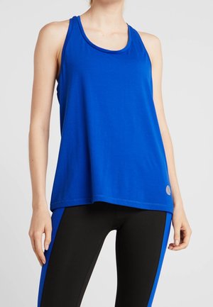 Kobieta w luźnym niebieskim topie bez rękawów i czarnych legginsach z niebieskimi paskami po bokach, stojąca na szarym tle.