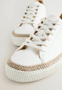 Sneakers bianchi con una parte superiore in sintetico liscio, rifinitura in juta intrecciata attorno alla punta e lacci bianchi piatti su una suola in gomma rigata.