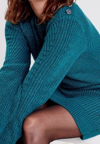 Pull tricoté bleu sarcelle avec texture côtelée, manches évasées et un bouton décoratif sur l'épaule. Porté par-dessus des collants noirs transparents.