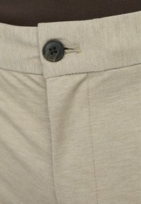 Béžové kalhoty s hladkou texturou, se černým knoflíkem a prošitím na pase a kapsách.