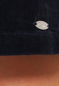 Navy corduroy-kangas, jossa on teksturoitu pinta. Siinä on hopeinen ovaali merkki, jossa lukee "Berliini", kiinnitetty tummalla langalla.