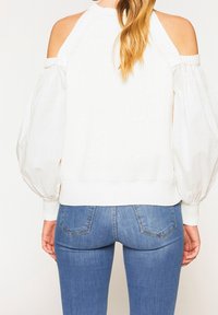 Pull en tricot blanc avec un design à épaule dégagée et des manches bouffantes. Porté avec un jean bleu ajusté, mettant en valeur une silhouette à la fois décontractée et structurée.