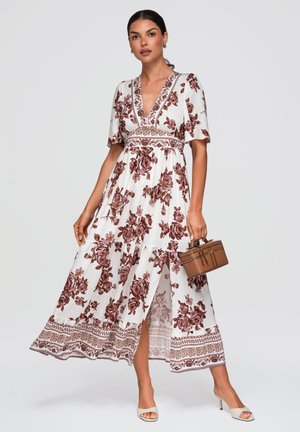 Femme portant une robe maxi fleurie blanche et marron à manches courtes, tenant un petit sac à main marron et portant des talons beiges ouverts à l'avant.
