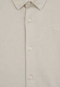 Világos bézs színű, texturált anyagú póló, gombos placket-tel és bal oldalon hímzett díszítéssel.