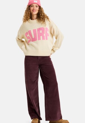 Pull en maille crème avec "SURF" en rose, associé à un pantalon large en velours côtelé bordeaux et des mocassins beiges. Bonnet rose sur la tête.