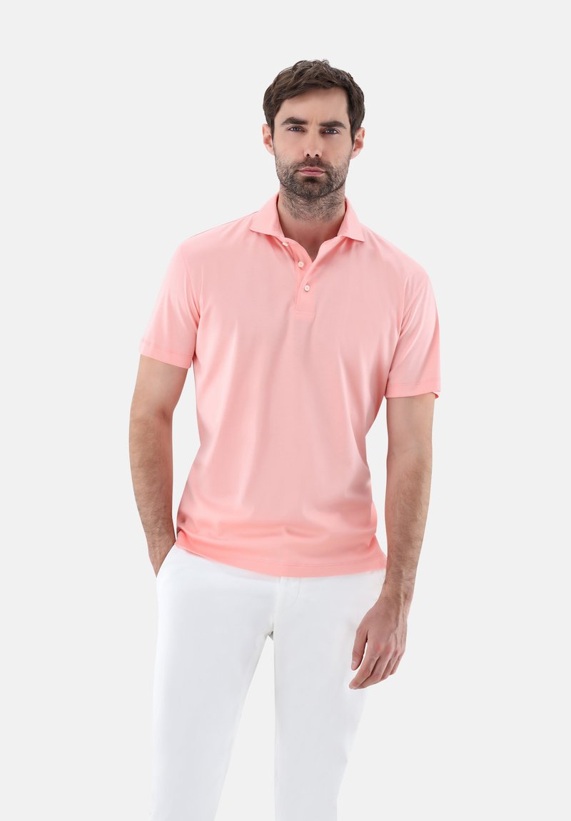 Rosa Polohemd mit kurzen Ärmeln, mit drei Knöpfen und weicher Textur, kombiniert mit weißen Hosen, Modell steht mit Händen in den Taschen.