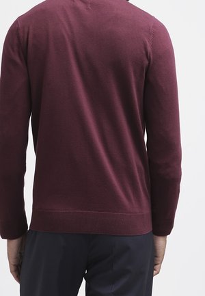 Homme portant un pull à manches longues ajusté bordeaux et un pantalon bleu marine, vu de dos devant un fond uni.