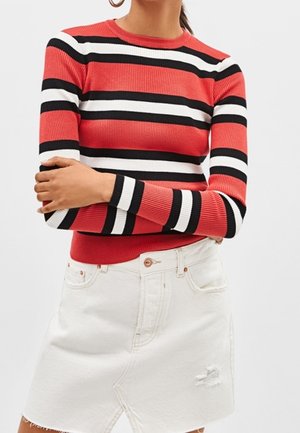 Persoon draagt een rood, zwart en wit horizontaal gestreept shirt met lange mouwen, gecombineerd met een witte denim rok met een klein scheurtje aan de linkerkant.