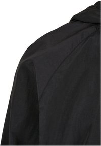 Gros plan d'une épaule de veste en tissu noir avec des coutures visibles et un col plié sur fond blanc.