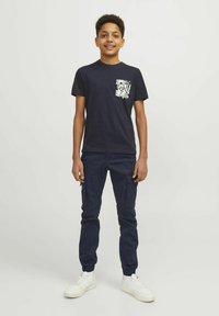 T-shirt à manches courtes bleu marine avec un design de poche florale blanche, associé à un pantalon cargo bleu marine et des baskets blanches. Coupe décontractée, texture lisse.