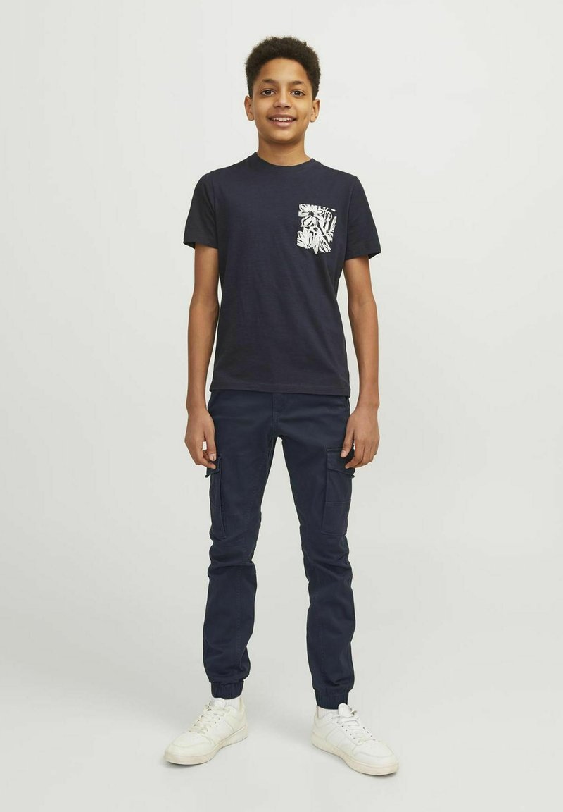 T-shirt à manches courtes bleu marine avec un design de poche florale blanche, associé à un pantalon cargo bleu marine et des baskets blanches. Coupe décontractée, texture lisse.