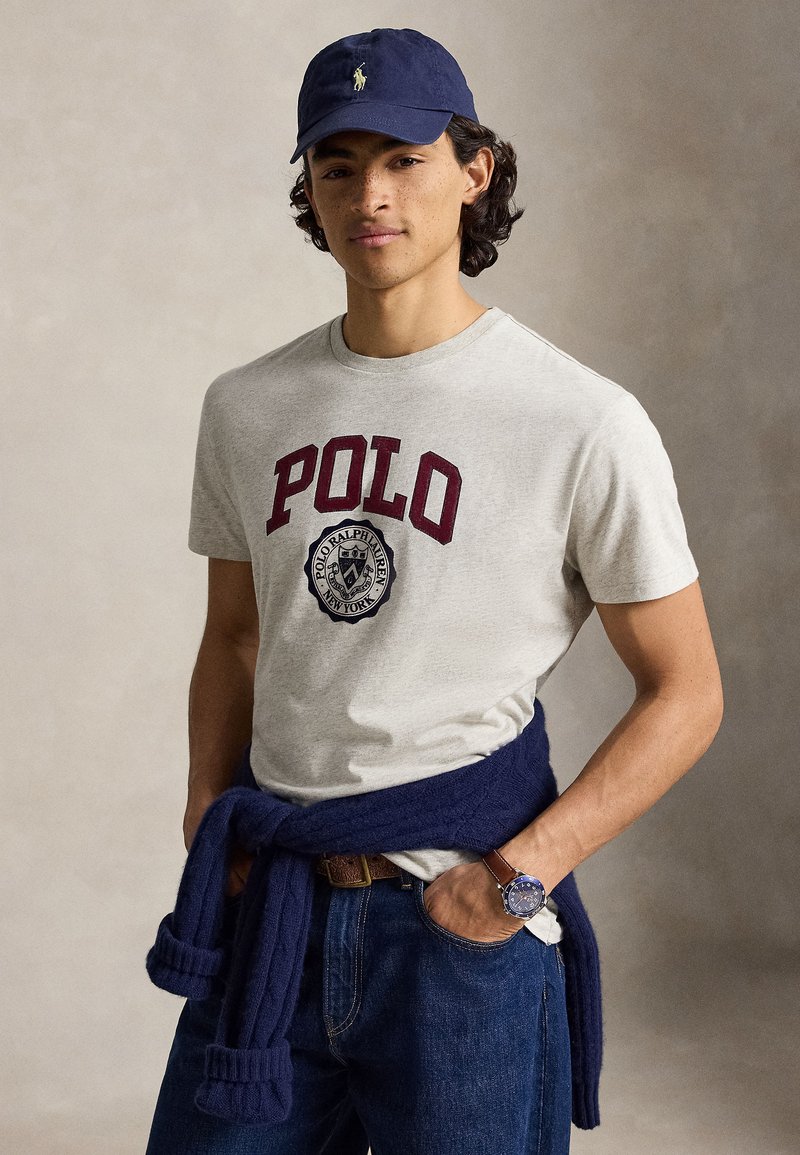 Νέος άνδρας που φοράει γκρι μπλουζάκι Polo Ralph Lauren, σκούφο ναυτικού χρώματος, σκούρο τζιν, με ένα πουλόβερ ναυτικού χρώματος δεμένο γύρω από τη μέση του και ένα ρολόι χειρός.