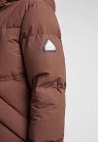 Chaqueta acolchada marrón con un parche de logo triangular en la parte superior del brazo. La tela parece suave con un ligero brillo y un diseño acolchado.