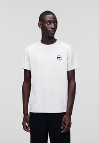 Tricou alb din bumbac cu guler rotund și mâneci scurte. Prezintă un logo grafic negru pe pieptul stâng. Design simplu, minimalist.