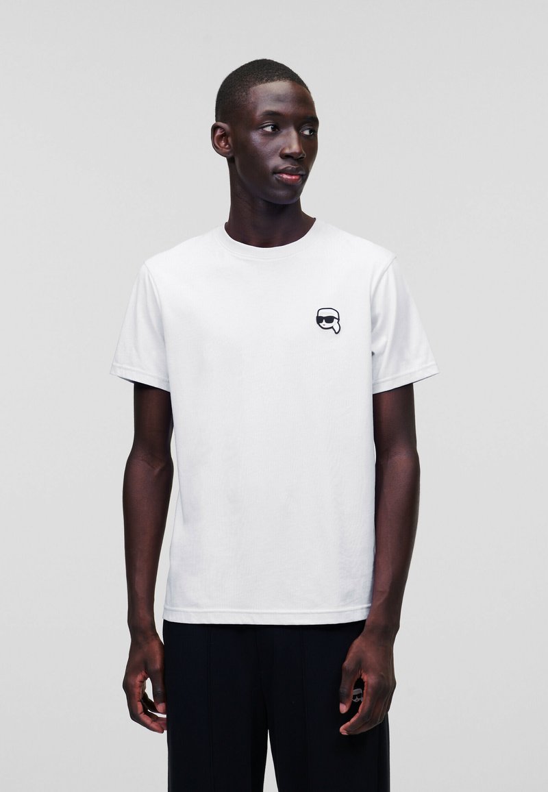 Tricou alb din bumbac cu guler rotund și mâneci scurte. Prezintă un logo grafic negru pe pieptul stâng. Design simplu, minimalist.