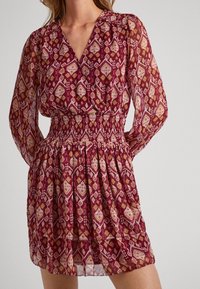 Rotes Kleid mit Muster und langen Ärmeln, das eine geraffte Taille und einen fließenden, geschichteten Rock aus leichtem, durchscheinendem Stoff aufweist.