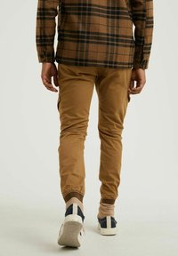 Brun rutmönstrad skjorta med en avslappnad passform, i kombination med ljusbruna slim-fit byxor. Byxorna har jogger-stil muddar och marinblå sneakers.