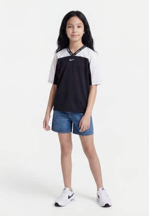 Ragazza con lunghi capelli neri che indossa una maglietta sportiva Nike bianca e nera, pantaloncini di jeans blu e scarpe da ginnastica Nike bianche, in piedi su sfondo bianco.