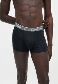 Torse masculin athlétique portant un boxer Nike noir avec une ceinture grise présentant des logos Nike blancs, debout avec un bras plié derrière le dos.
