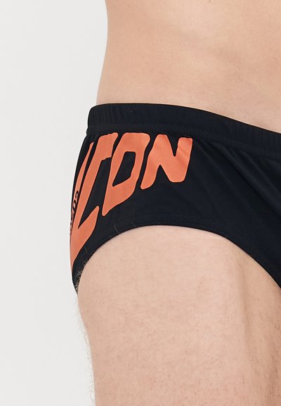 Mann trägt schwarze Badebriefs mit großen orangefarbenen "ICON"-Text an der Seite, der einen Teil des Oberkörpers und des Oberschenkels zeigt.