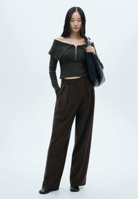 Donkergrijze off-shoulder top met ritsdetail, gecombineerd met bruine wijde broek. Het model draagt een zwarte schoudertas en heeft zwarte schoenen aan.
