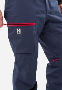 Pantalons de ski imperméables bleu marine avec poches zippées rouges et logo Millet. Présente une texture lisse et une coupe fuselée.