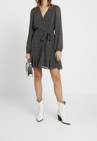 Svart omlottklänning med långa, genomskinliga ärmar, prickigt mönster, knyt i midjan och kjol med utsvängd fåll. Matchas med vita cowboystövlar med klack och en silverfärgad väska.