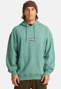 Teal hoodie with a front pocket, drawstring hood, and ribbed cuffs. Features a small embroidered logo on the chest. Soft, relaxed fit.