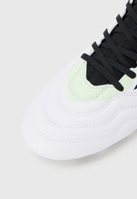 Hvid sneaker med tekstureret overflade, sorte snørebånd og grønne accenter. Rundt tådesign og lagdelte syningsdetaljer.