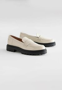 Mocassini in pelle verniciata beige chiaro con una larga suola in gomma nera, caratterizzati da un dettaglio cucito e un design flat classico.