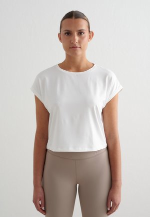 Kobieta stojąca zwrócona przodem, ubrana w białą, krótką koszulkę i wysokiej talii legginsy w kolorze taupe na jednolitym tle.