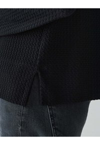 Schwarzer strukturierter Pullover mit einem Waffelstrickdesign, ausgestattet mit seitlichen Schlitzen und einer lockeren Passform. Kombiniert mit dunkelgrauen Jeans.