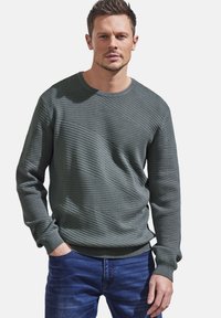 Grüner, strukturierter Pullover mit Rundhalsausschnitt und diagonaler Rippenstruktur, kombiniert mit blauen Jeans; lässiges Design.