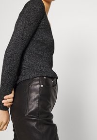 Nainen, jolla on päällään musta kimaltava pitkähihais toppi, joka on tettu korkeavyötäröisten mustien nahkahousujen sisään, poseeraa vaaleaa taustaa vasten.