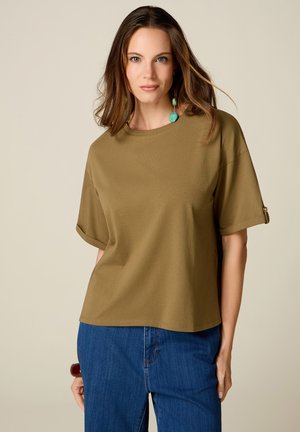 Femme aux cheveux bruns mi-longs, portant un haut vert olive à manches courtes et un jean bleu, debout devant un fond beige uni.