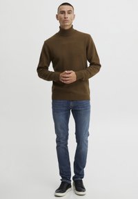 Rudas turtleneck megztinis minkštu mezgimu, derinamas su mėlynomis džinsomis ir juodais sportbačiais. Modelis stovi tiesiai, rankos sukryžiuotos priešais.
