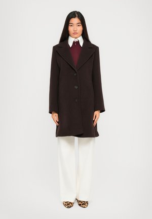 Woman standing wearing a dark brown coat over a burgundy top with white collar, white wide-leg pants, and leopard print shoes on a white background.