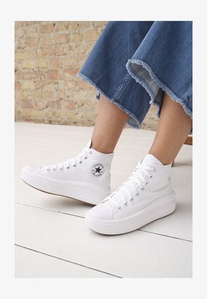 White high-top sneakers made of canvas with a rubber sole, featuring silver eyelets, a round logo patch, and a textured platform sole.