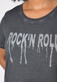 Grafitově šedé tričko s kulatým výstřihem, zdobené stříbrným glitrovým potiskem "ROCK'N ROLL", který stéká dolů, na měkkém, strukturovaném materiálu.