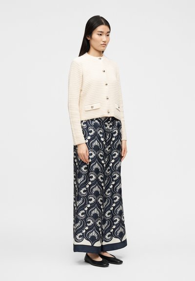 Γυναίκα που φοράει κρεμ υφασμένο ζακέτα με κουμπιά και φαρδύ παντελόνι σε ναυτικό μπλε με λευκό ανθικό μοτίβο, στέκεται με τα χέρια στο πλάι.