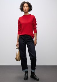 Maglione rosso lavorato a maglia con maniche lunghe, jeans blu scuro con orli risvolti e stivaletti neri in pelle; tiene una borsa a tracolla beige.